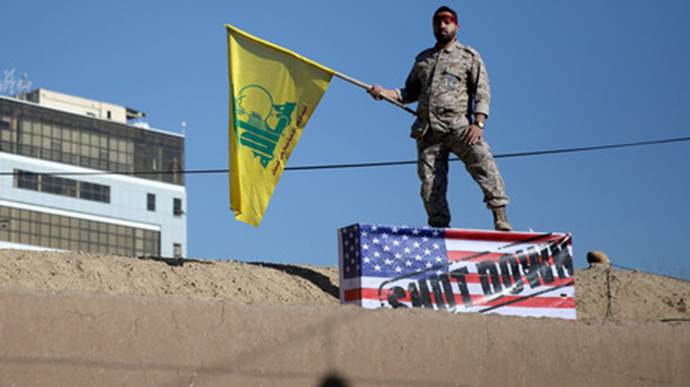 Eine iranische Wache hlt eine Hisbollah-Flagge whrend des Trauerzuges bei der Beerdigung von Soleimani in Kerman und steht dabei auf einer US-Flagge. Auf dieser steht: 